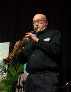 Phil Mershon plays a gold saxophone on stage in front of dark curtains.